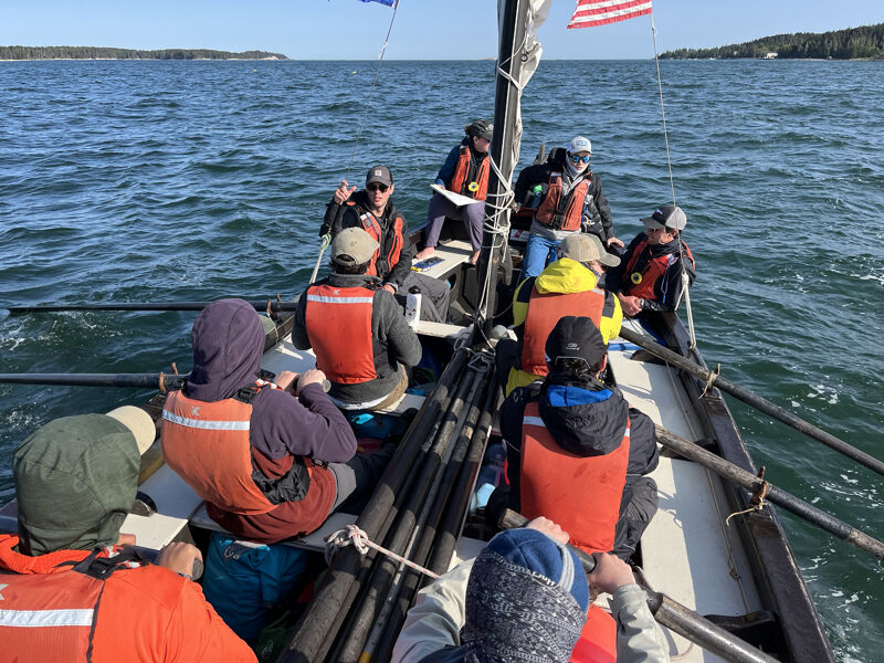 The image shows a group of people on a boat, likely engaged in a rowing activity. They are wearing life jackets, suggesting a focus on safety. The presence of flags and the open water in the background indicate an outdoor setting. The boat appears to be a traditional rowboat, and the individuals seem to be participating in a recreational or training exercise.